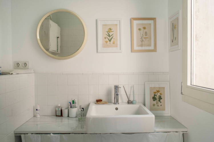 Imagen parcial de un baño donde se ve un lavabo blanco apoyado en una encimera. En la pared hay un espejo redondo y 2 cuadros decorativos